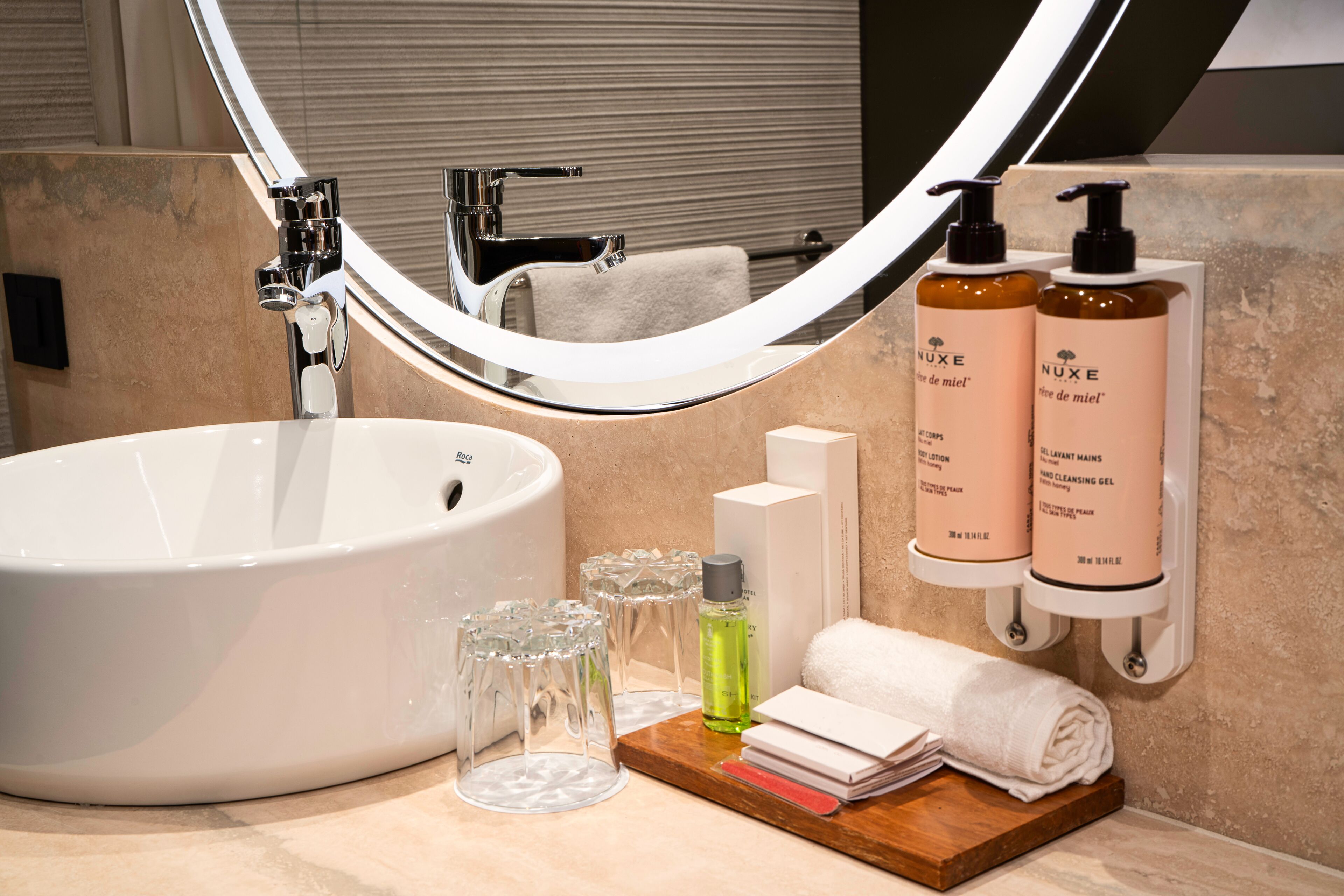 Un baño con un lavabo grande de color blanco, un espejo y dos botellas de productos Nuxe para el cuidado de la piel.