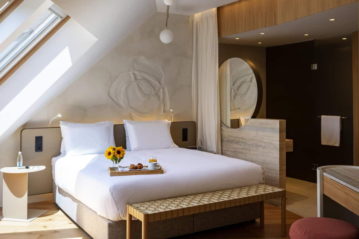 A modern hotel room with a large bed, a bedside table with a tray of breakfast, a mirror, and a view of the outdoors.