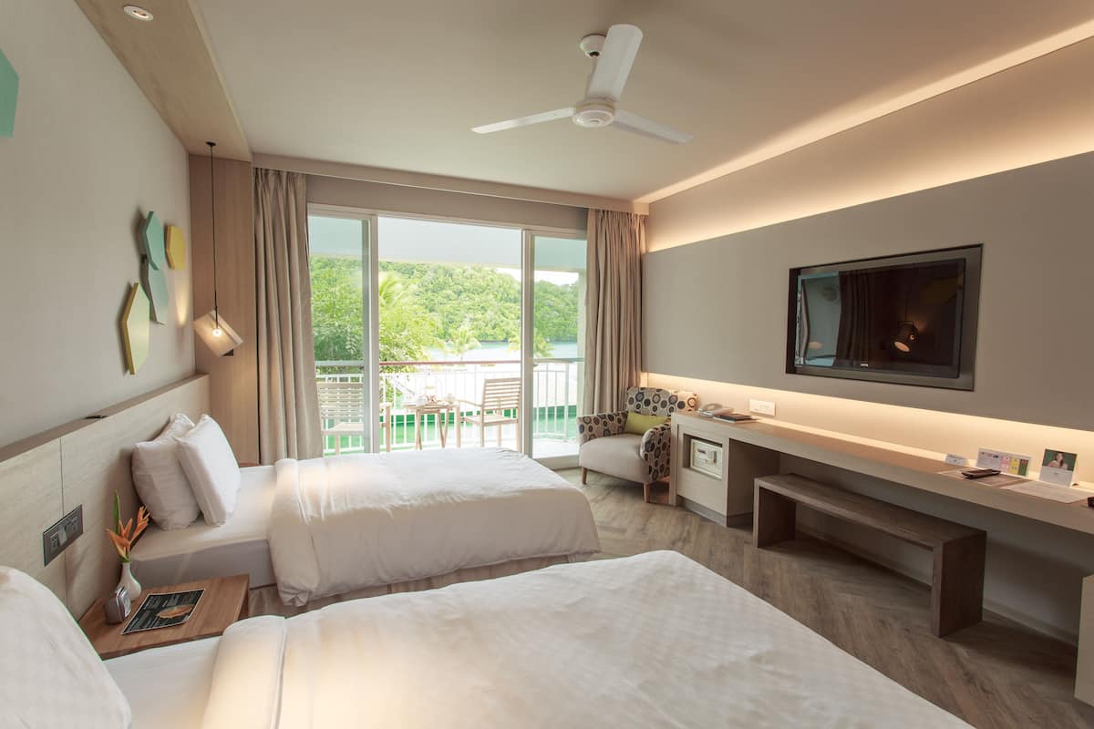 Une chambre d’hôtel avec deux lits, un bureau, une télévision et un balcon donnant sur un espace verdoyant.