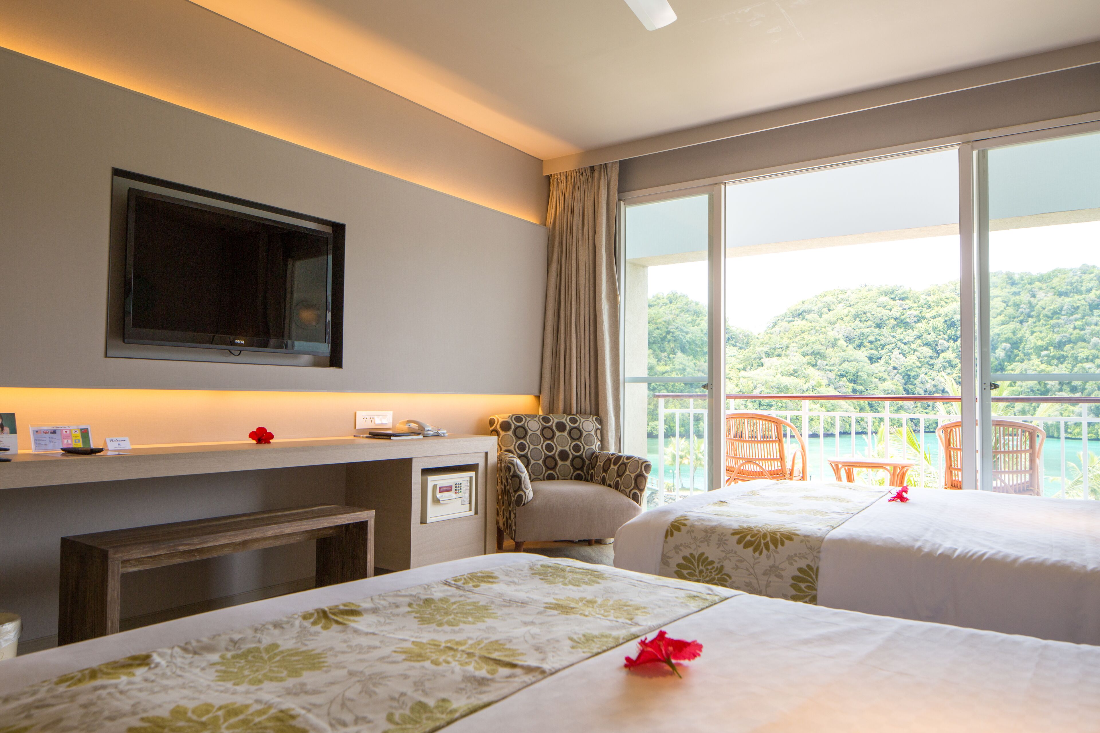 Habitación de hotel con dos camas, televisión, escritorio y balcón con sillas.