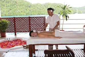 Une séance de massage en plein air, sur une terrasse en bois, avec vue sur une colline boisée et un plan d’eau.