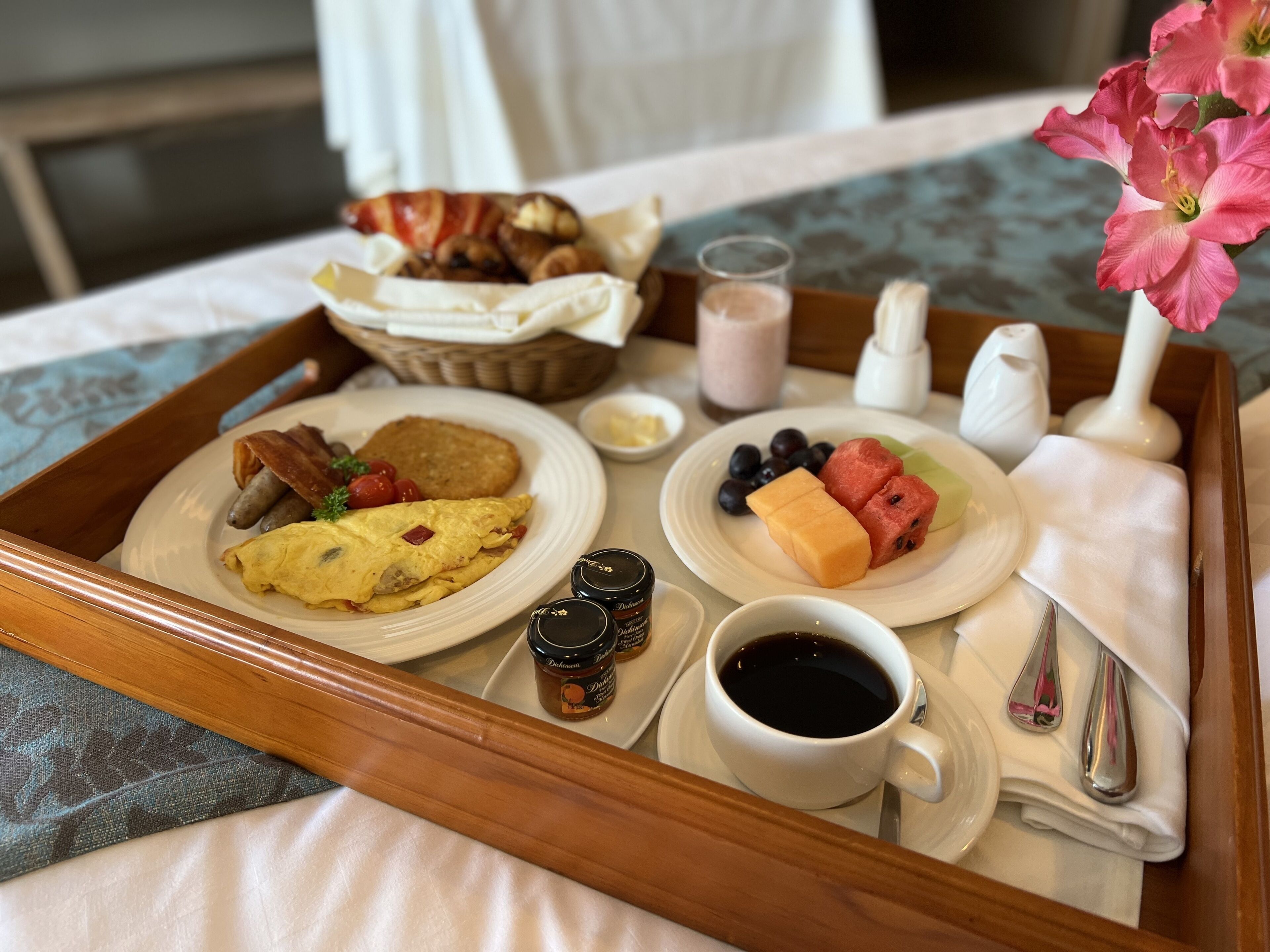 Bandeja de desayuno con huevos, salchichas, tostadas y fruta sobre una cama con un cojín estampado con motivos florales.
