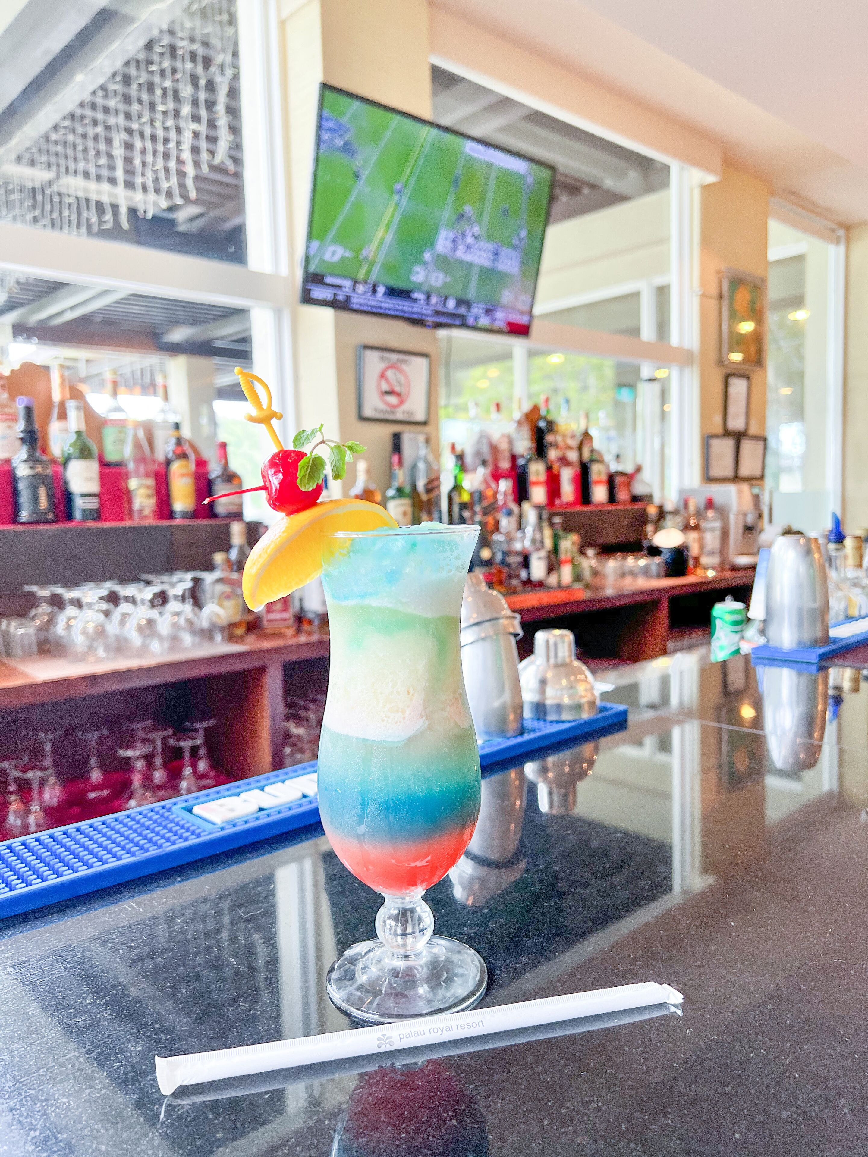 Zona de bar con un cóctel de colores, una pantalla de televisión con deportes y una variedad de botellas y vasos.