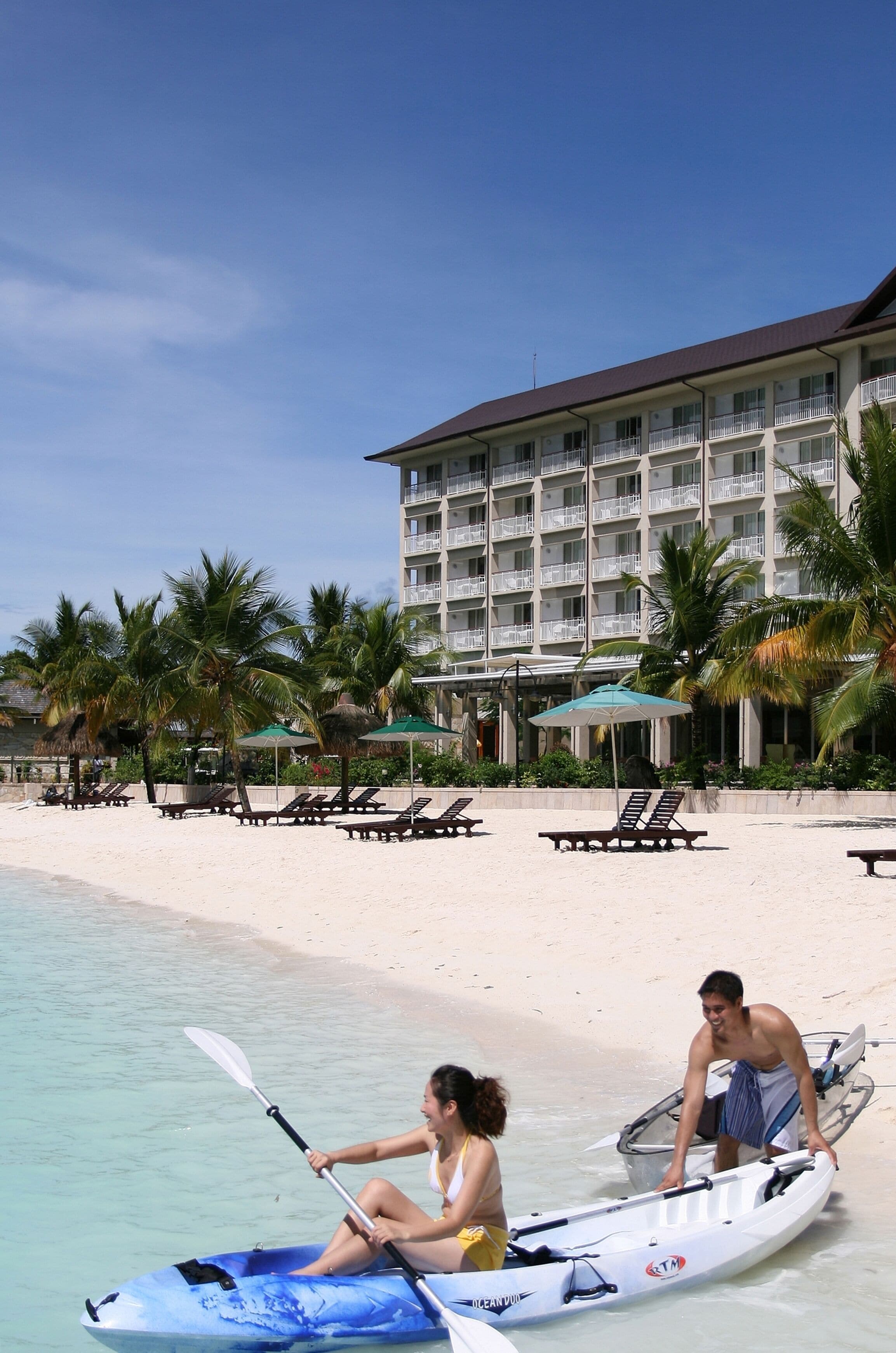 Un resort frente al mar con un edificio grande, palmeras y sillas de descanso.