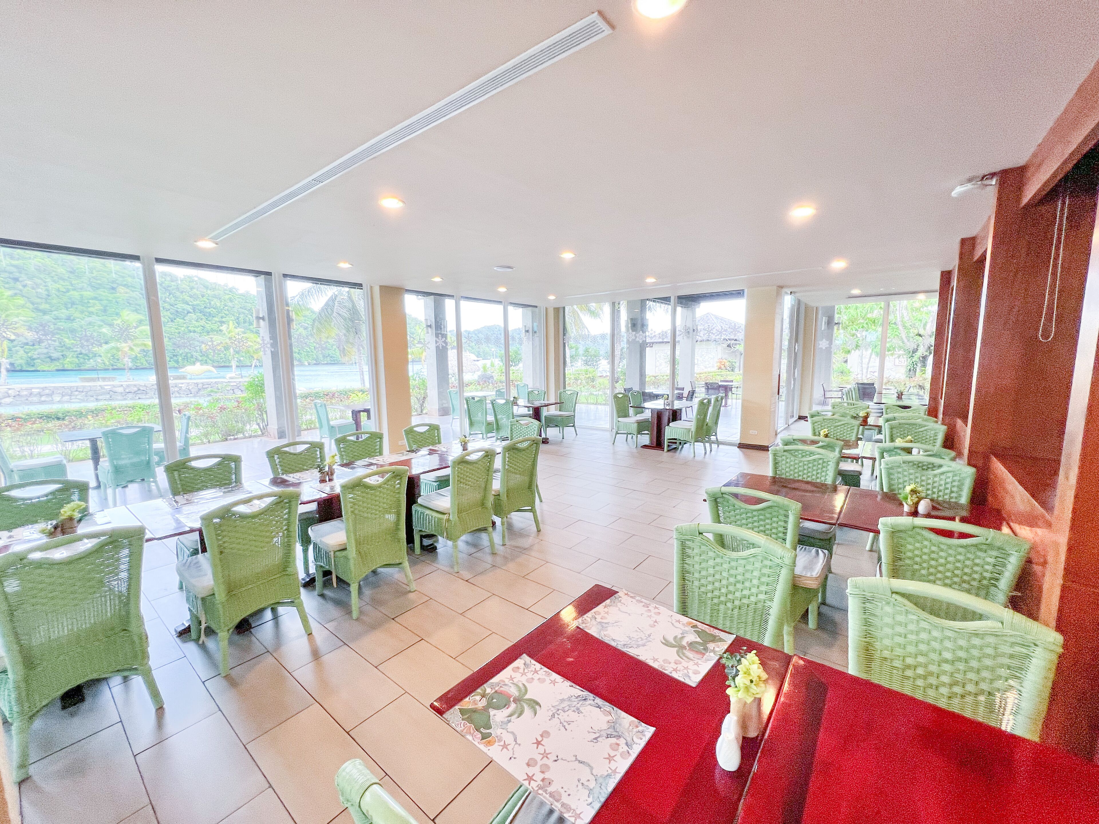 A spacious indoor dining area with green wicker chairs and tables, large windows offering a view of the outdoors.