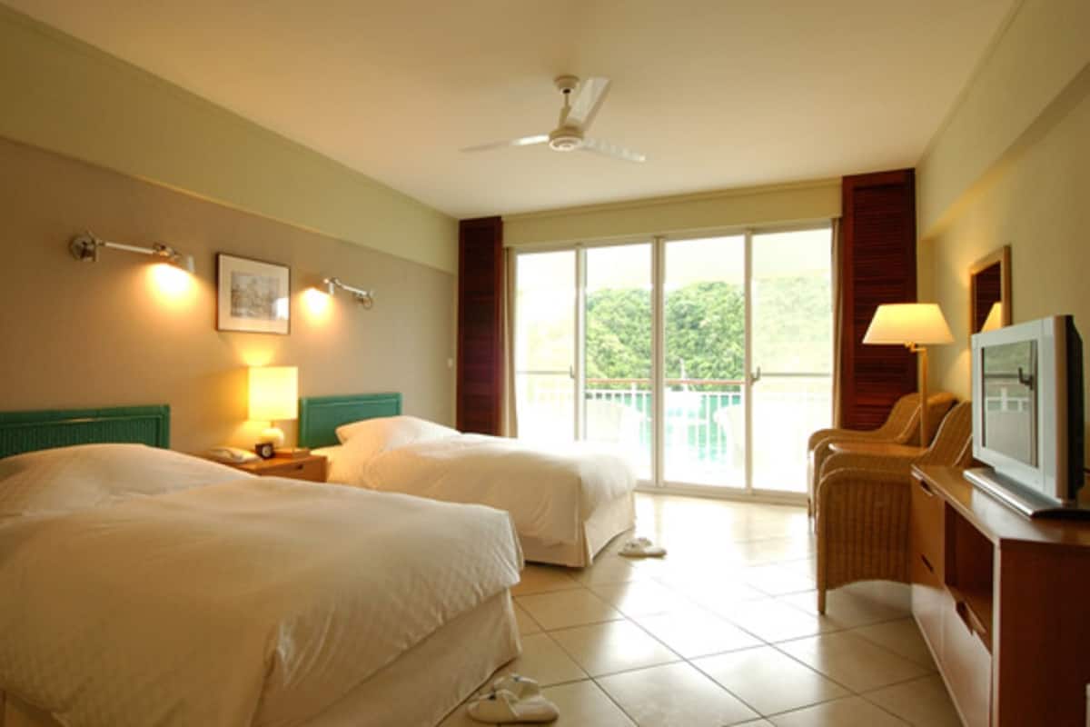 Une chambre d’hôtel avec deux lits, une télévision, un bureau avec une lampe et un balcon donnant sur un espace verdoyant.