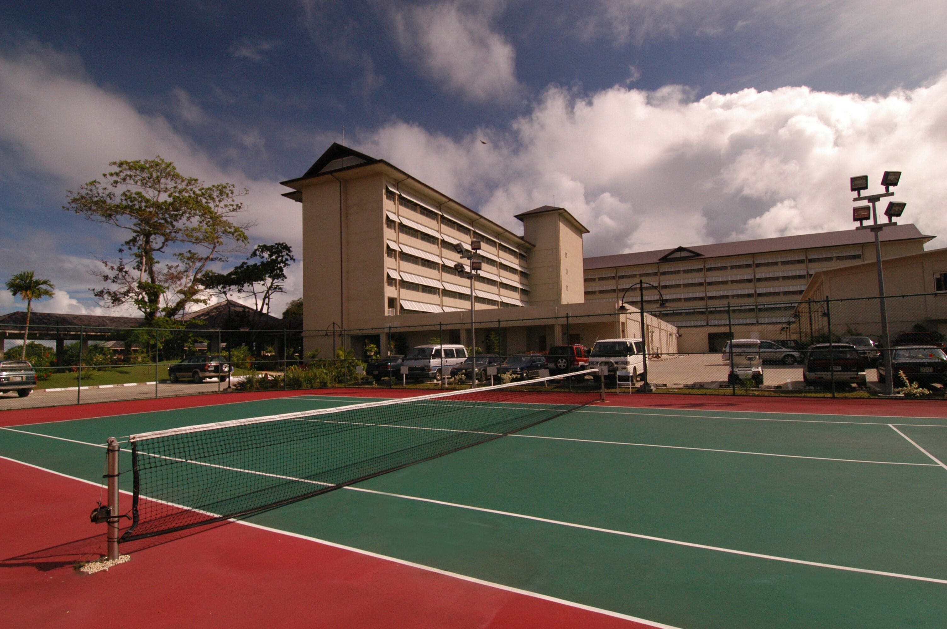 An outdoor tennis court with a net, surrounded by a parking area and a building with multiple floors and balconies.