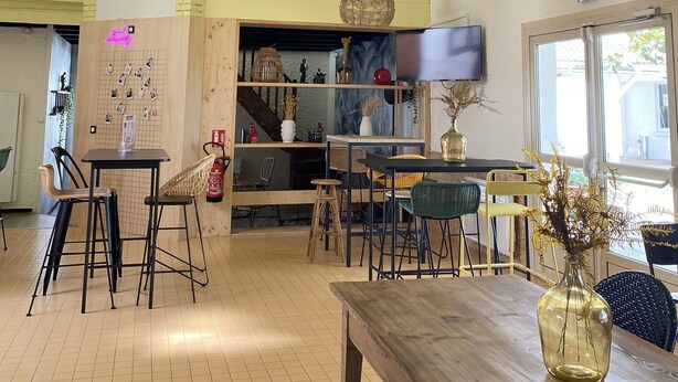 Ein geräumiges Café mit hohen Decken, freiliegenden Balken und hängenden Korbstühlenlampen. Tische und Stühle sind zum Sitzen angeordnet, im Hintergrund ist eine Theke erkennbar.
