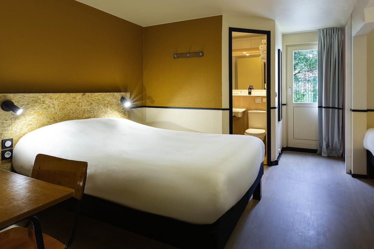 Habitación de hotel con cama, escritorio, silla, mesita de noche, espejo y ventana con cortinas.