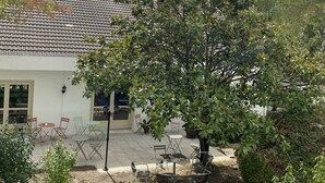 Un edificio con techo de tejas, un patio con mobiliario exterior y un árbol en primer plano.