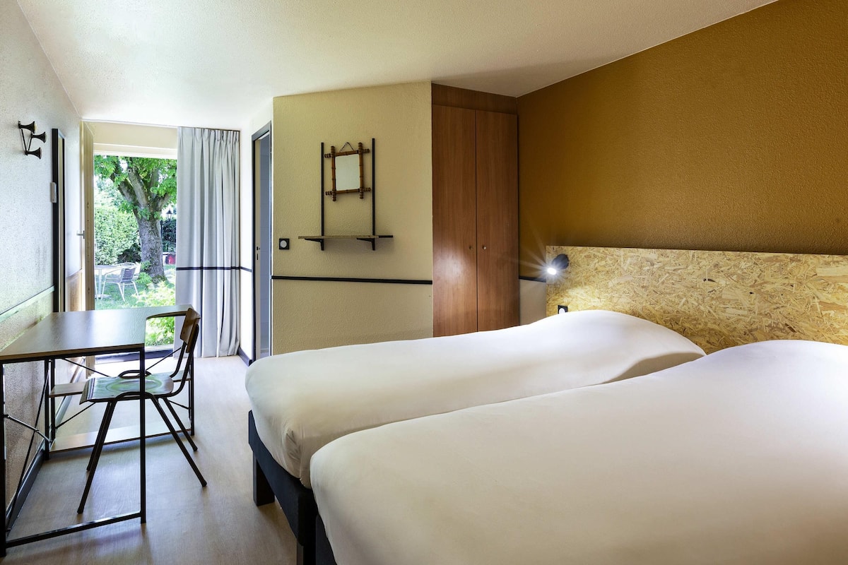 Habitación de hotel con dos camas, un escritorio, una silla, una ventana con cortinas y una mesita.