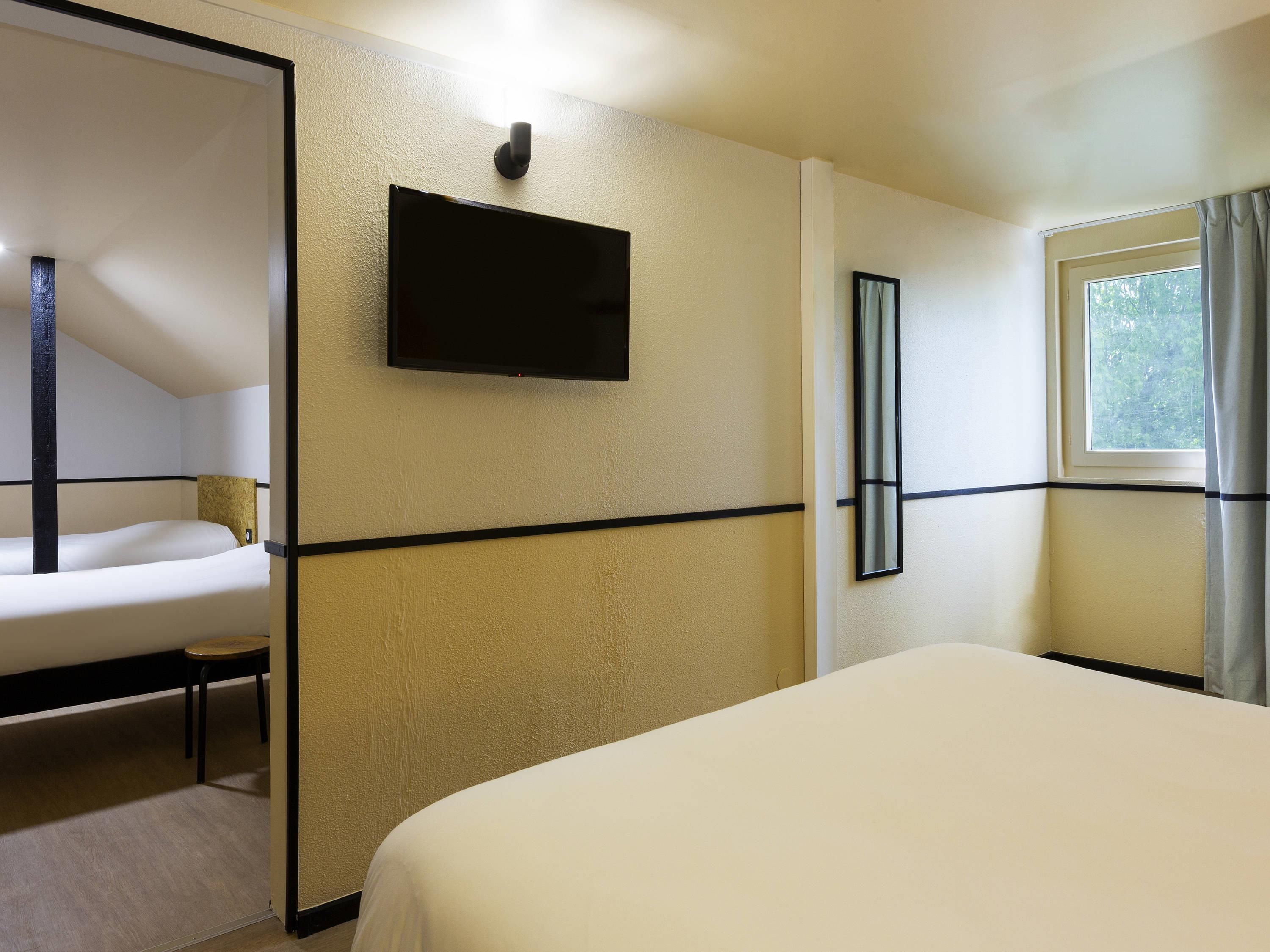 A hotel room with a bed, a television mounted on the wall, a window with curtains, and a small table with a chair.