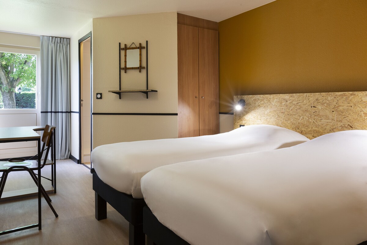 Habitación de hotel con dos camas, un escritorio con silla, un armario y una ventana con cortinas.