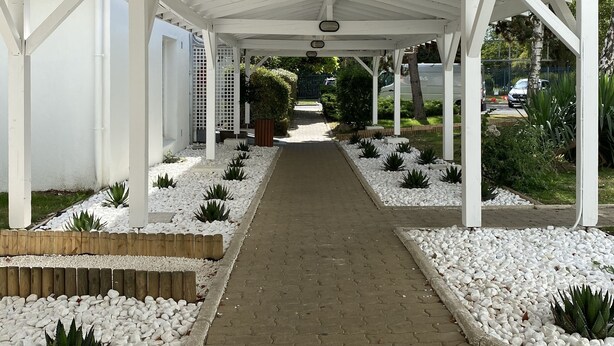 Un paseo techado con pilares blancos y un sendero de ladrillo, rodeado de guijarros blancos y pequeñas plantas.