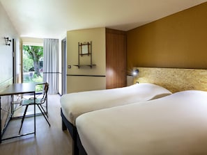 Ein Hotelzimmer mit zwei Betten, einem Schreibtisch, einem Stuhl, einem Fenster mit Vorhängen und einem kleinen Tisch.