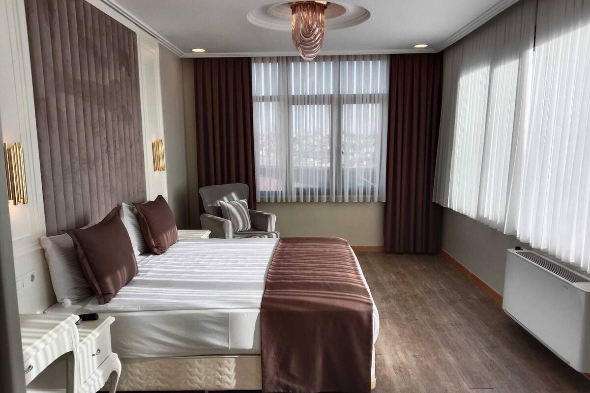 Quarto de hotel moderno com uma cama grande, uma poltrona, uma escrivaninha e uma janela com cortinas.