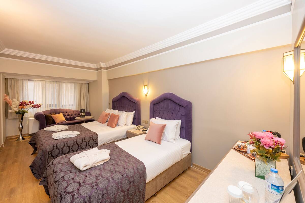 Quarto de hotel com duas camas, um sofá, uma escrivaninha com um vaso de flores e um espelho.