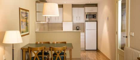Una cocina compacta con armarios blancos, un microondas y un refrigerador. Hay una mesa de comedor con sillas y una lámpara de pie.