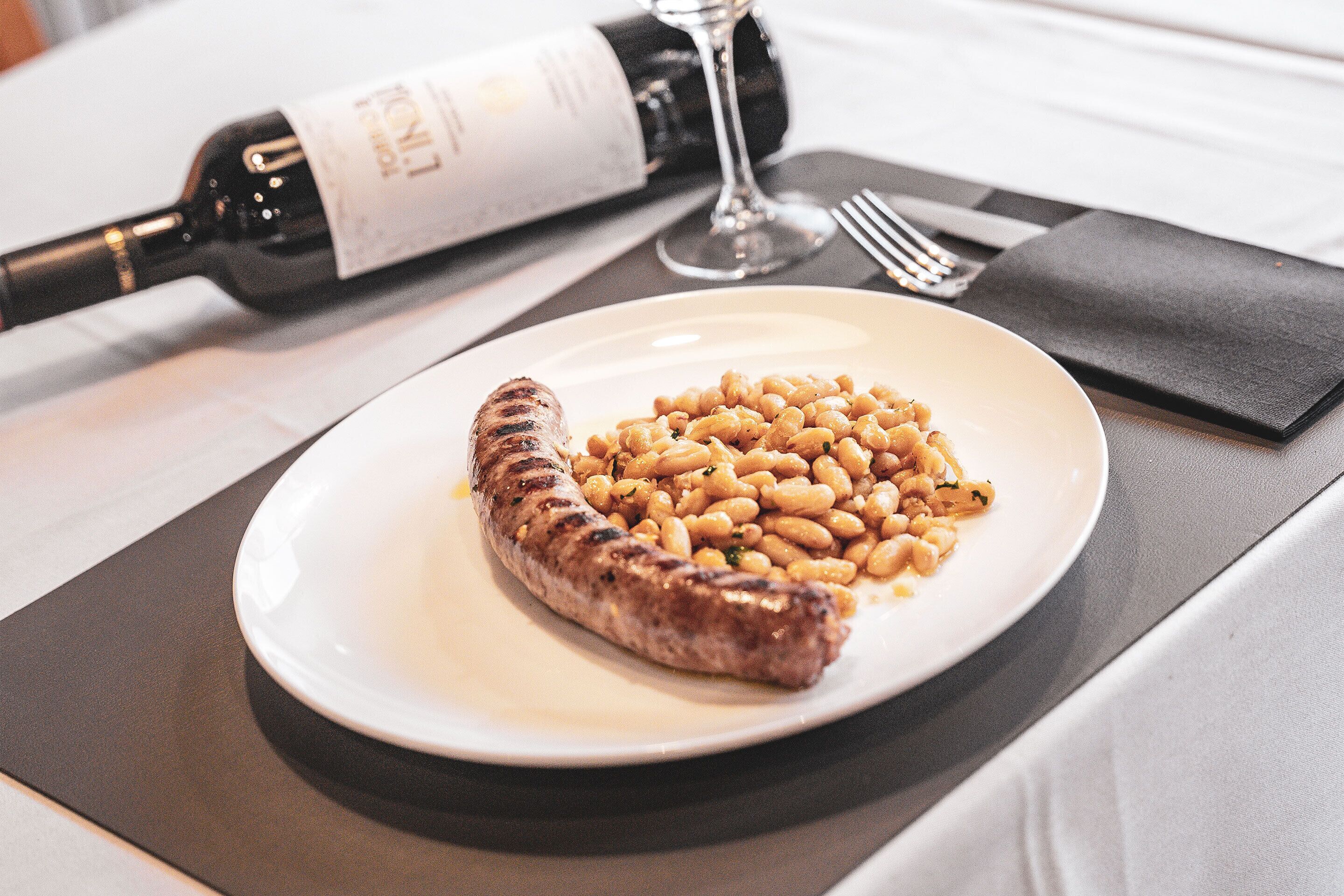 Une assiette avec une saucisse grillée et des haricots, une bouteille de vin et un verre de vin sur une table.