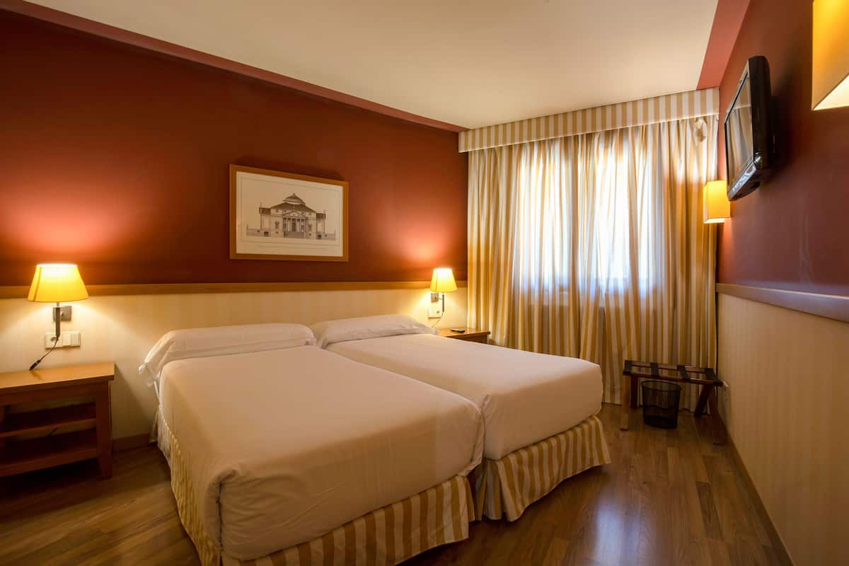 Habitación de hotel con dos camas, televisor montado en la pared, un cuadro enmarcado y una ventana con cortinas.