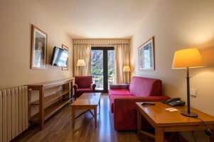 A hotel room with a wooden floor, a red sofa, a wooden table, a TV mounted on the wall, and a window with curtains.