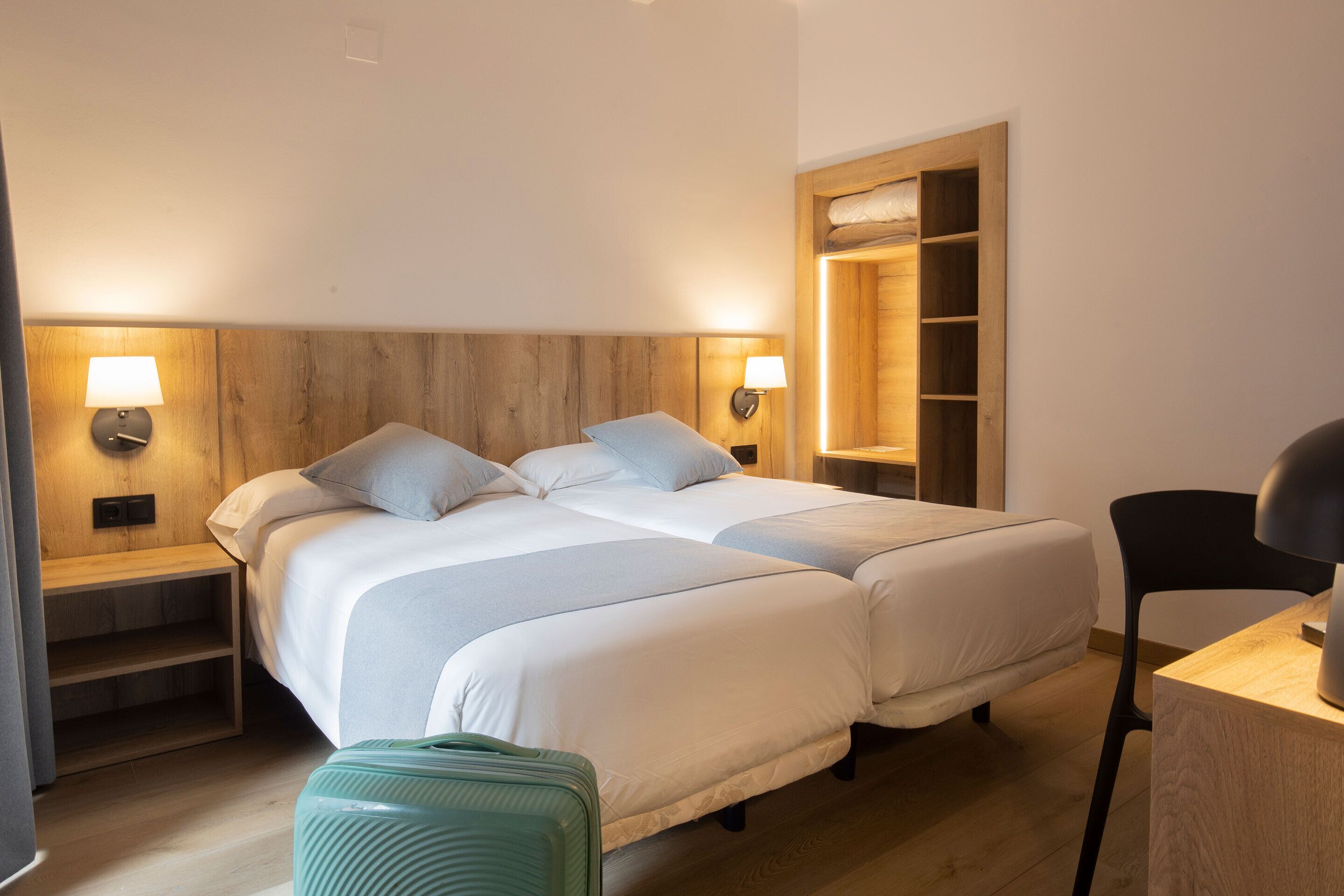 Hotellihuone, jossa on kaksi sänkyä, yöpöydällä olevat lamput, puinen sängynpääty, työpöytä tuolilla ja matkalaukku.