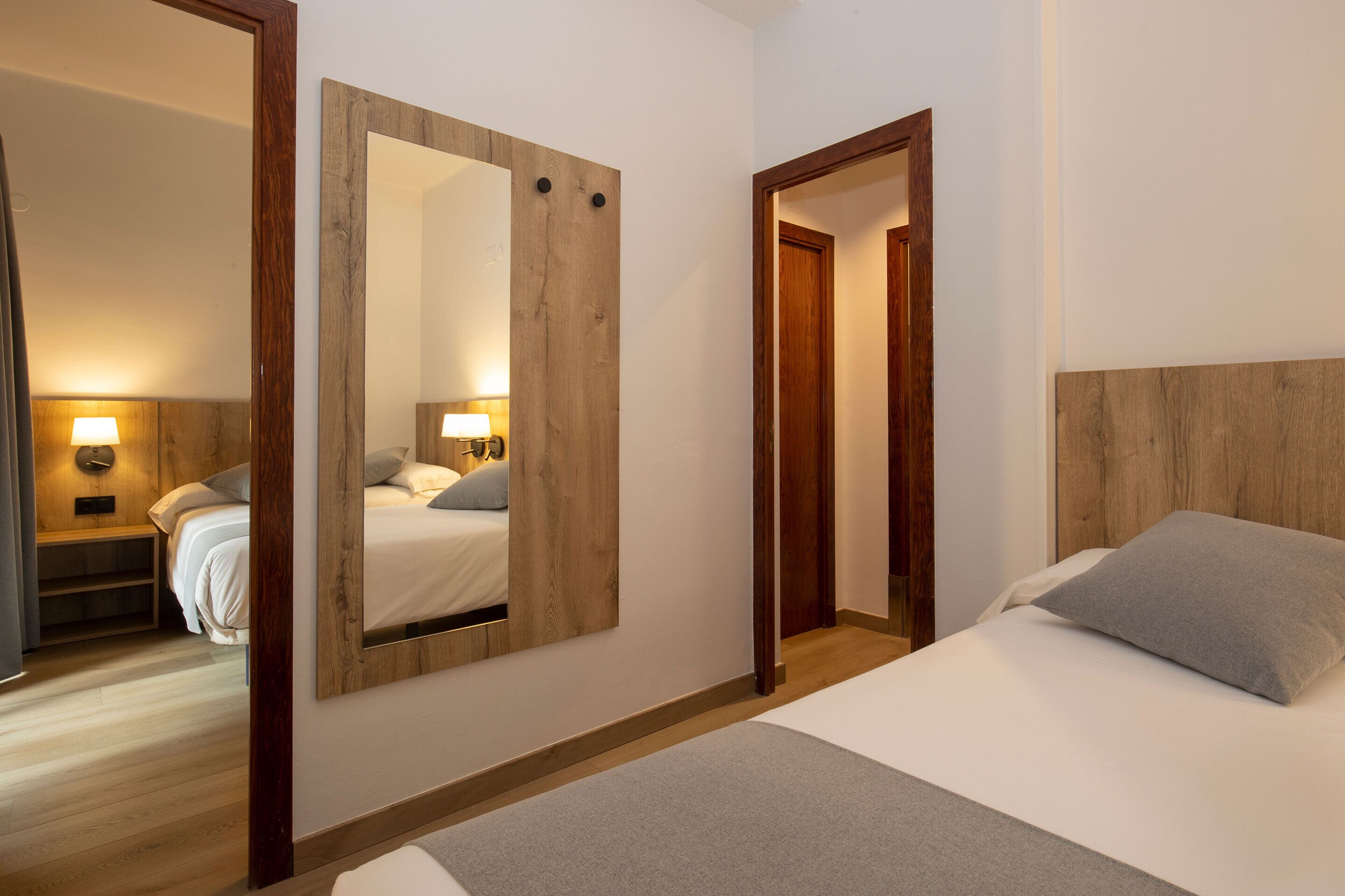 Una habitació d'hotel amb un llit, un panell de capçalera de fusta, un mirall gran i una tauleta de nit amb una làmpada.