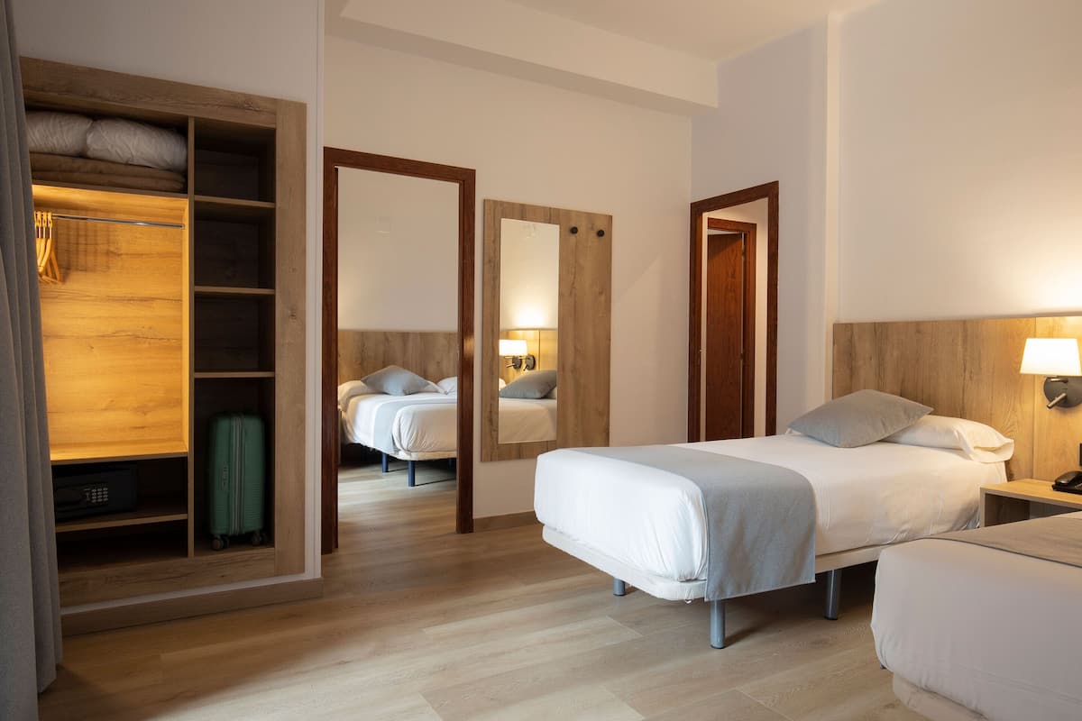 A hotel room with a wooden wardrobe, a bed with a white bedspread, a bedside table with a lamp, and a mirror on the wall.