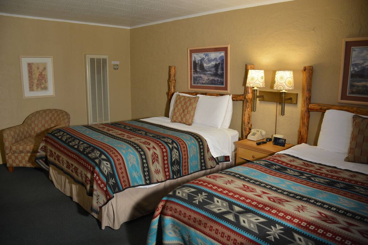 A hotel room with two beds, a chair, a nightstand with a phone and lamp, and a framed picture on the wall.
