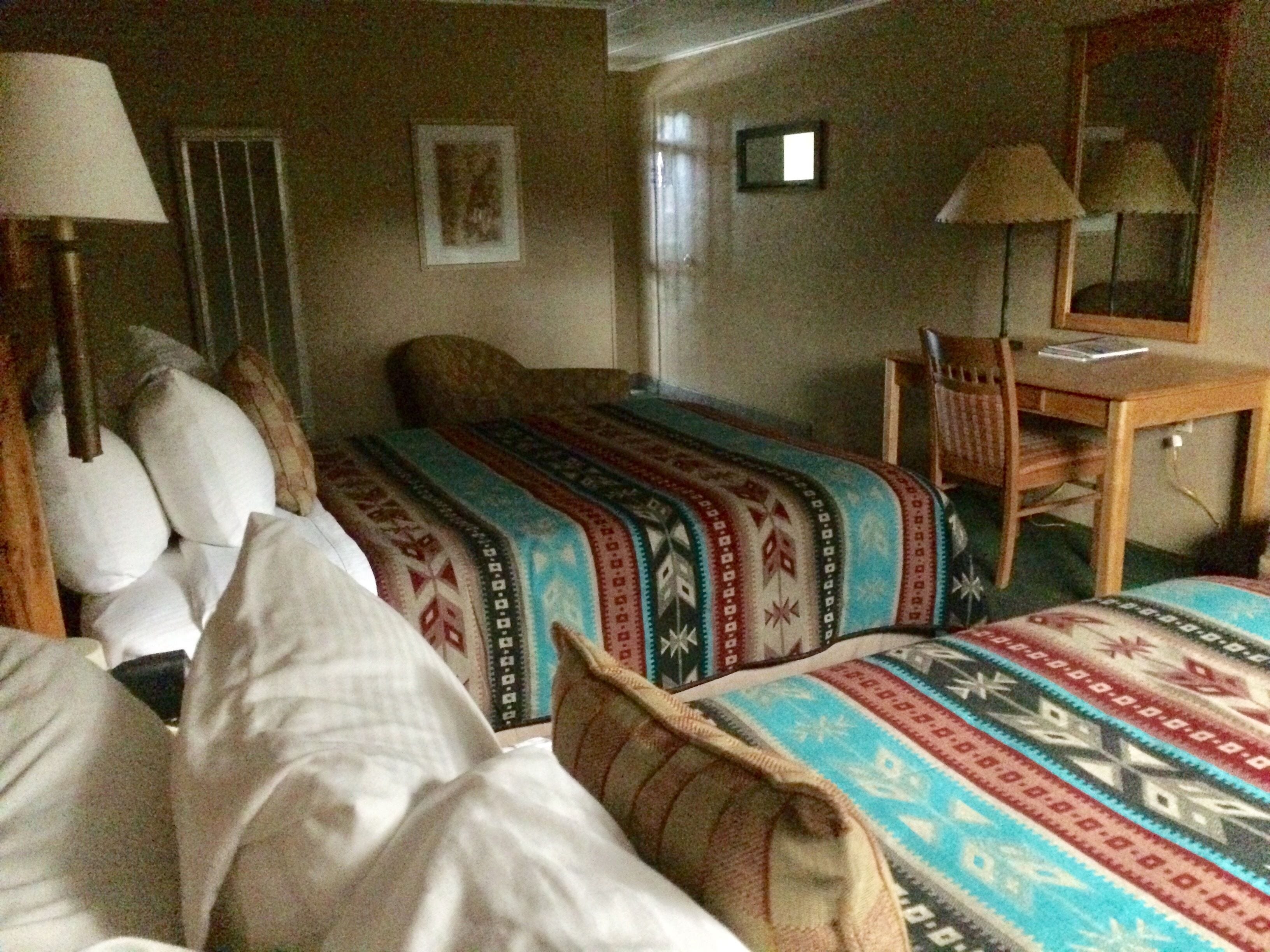 A hotel room with a bed, a desk, and a chair. There is a lamp on the desk and a picture on the wall.