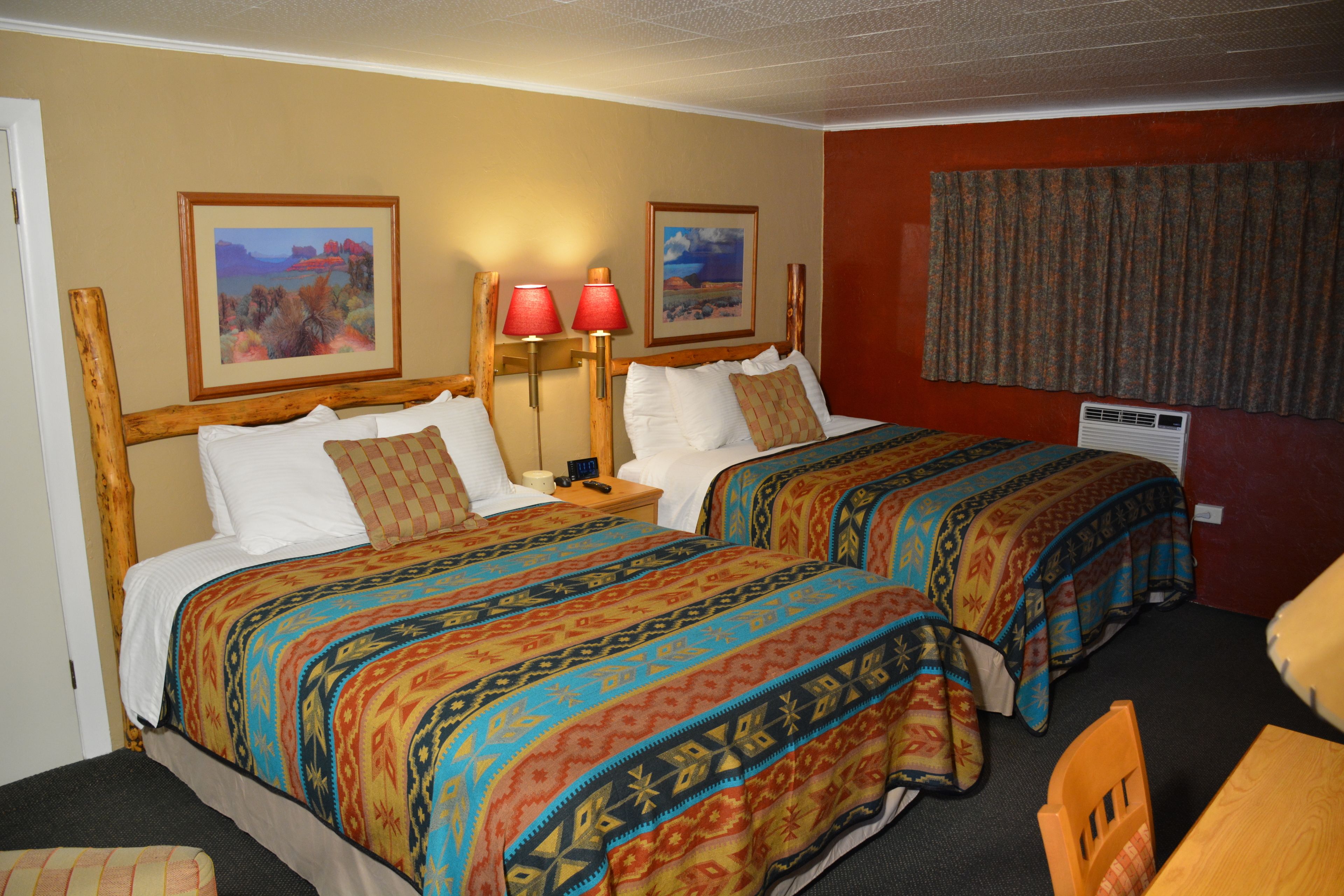 Une chambre d’hôtel avec deux lits, chacun recouvert d’une courtepointe aux couleurs vives, une table de chevet avec une lampe et deux tableaux encadrés accrochés au mur.