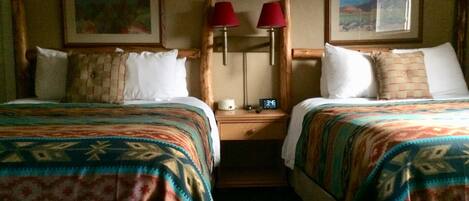 A hotel room with two beds, each with patterned bedding, a nightstand with a clock, and two red lamps.