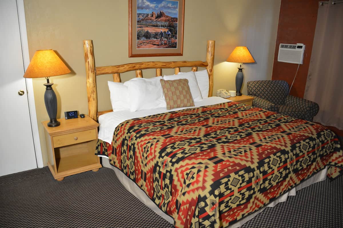 A hotel room with a wooden bed, two bedside lamps, a nightstand, a chair, and a painting on the wall.