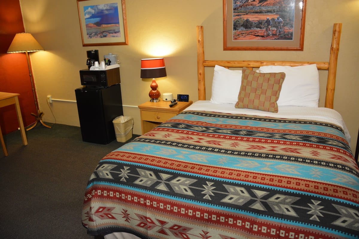 A hotel room with a bed, a nightstand, a lamp, a coffee maker, a remote control, and a picture on the wall.