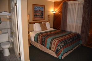 A hotel room with a bed, a painting on the wall, a lamp, and a bathroom visible through an open door.