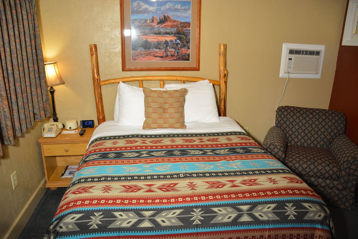 A hotel room with a bed, a nightstand, a lamp, a chair, a painting on the wall, and an air conditioning unit.