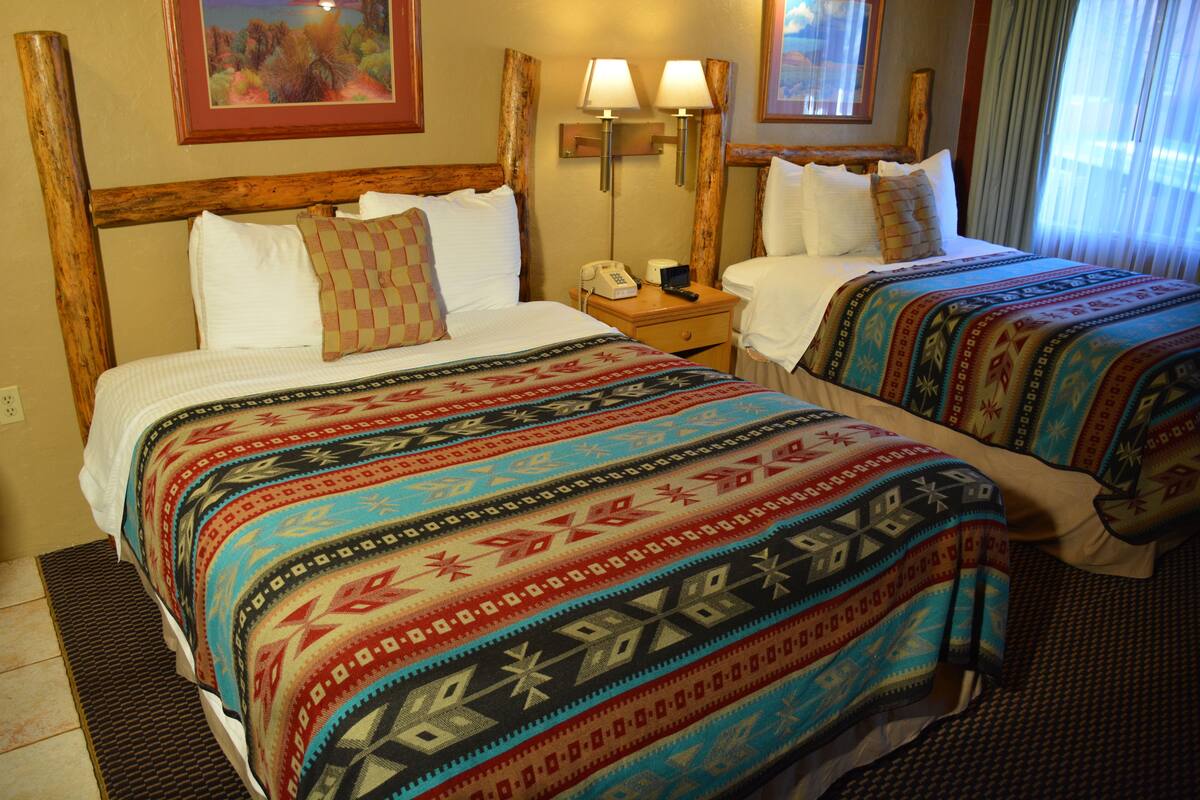 A hotel room with two beds, each with a patterned bedspread, a wooden headboard, a nightstand with a telephone, and wall-mounted lamps.
