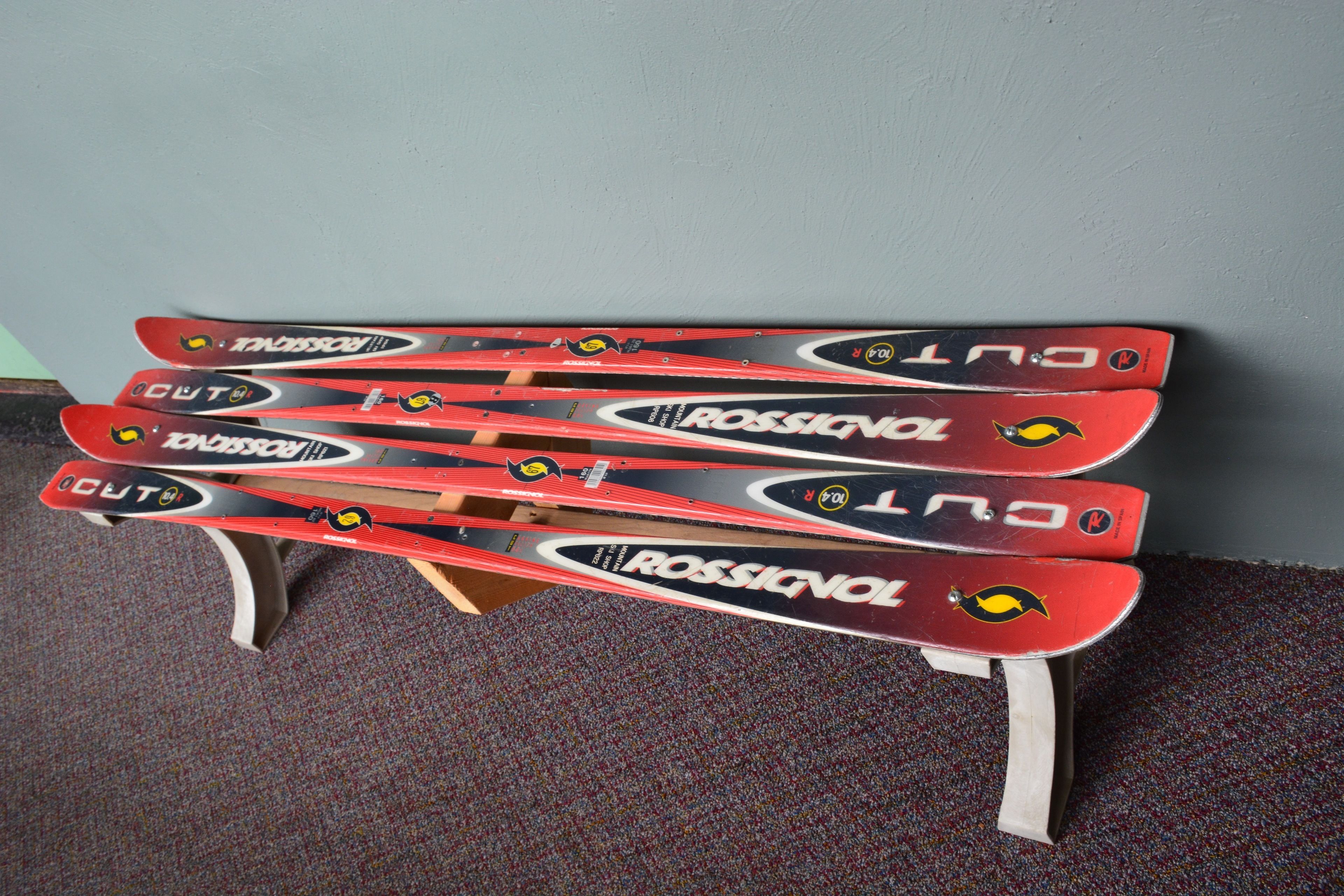 Une paire de skis Rossignol, rouges et noirs, est présentée sur un présentoir en bois, le tout adossé à un mur gris.