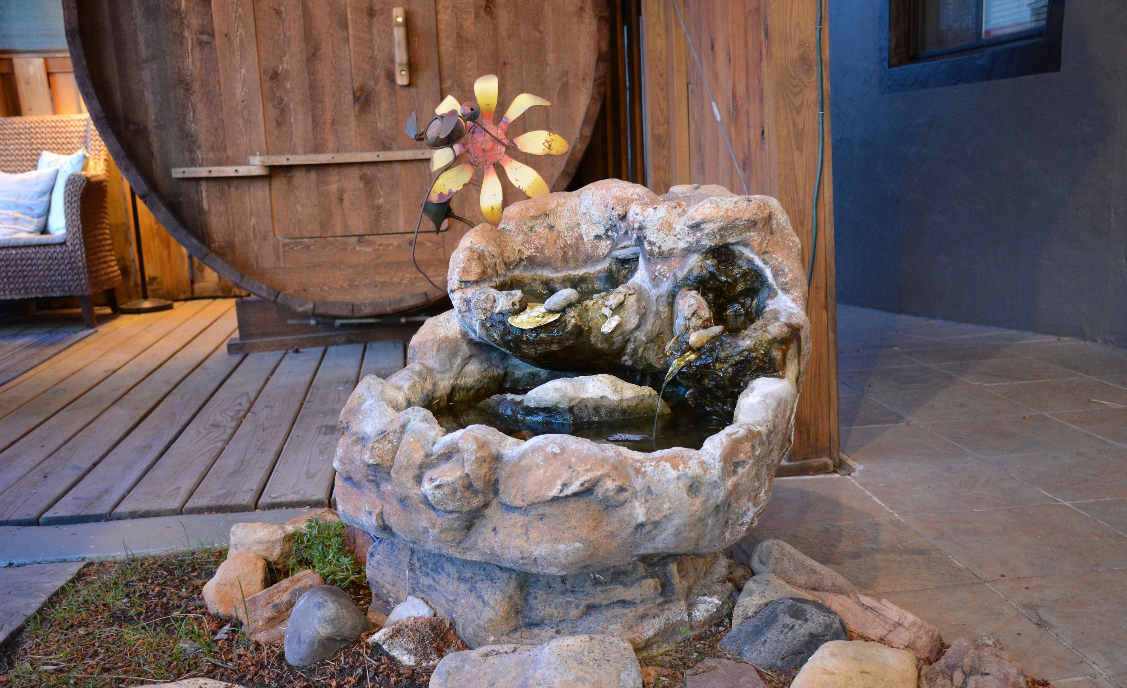 Une fontaine en pierre où l’eau jaillit, entourée de rochers et d’herbe, devant une porte en bois et une chaise en osier.
