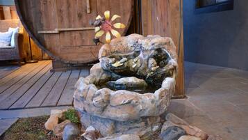 Una fuente de piedra con agua corriendo, rodeada de rocas y césped, frente a una puerta de madera y un sillón de mimbre.