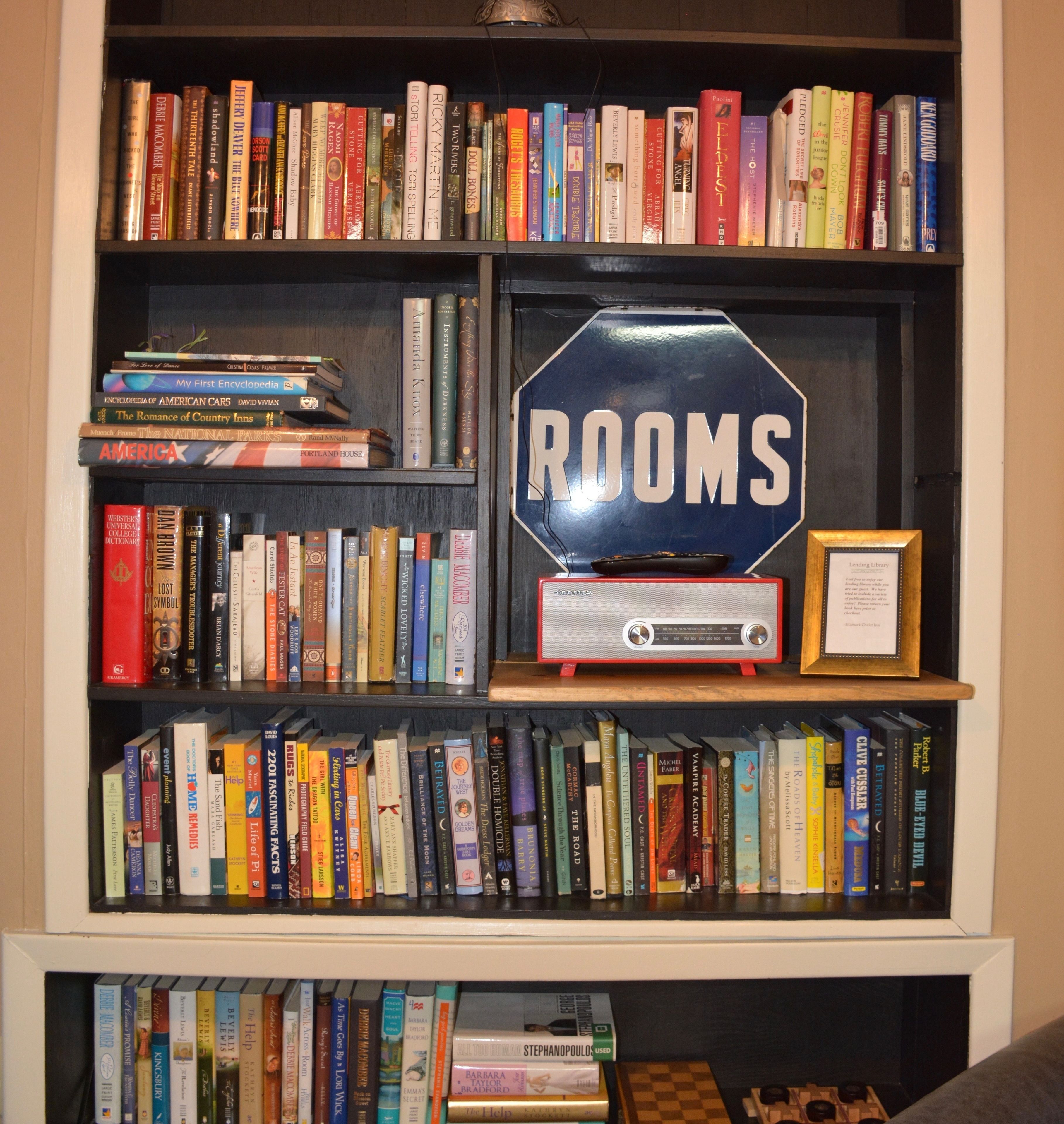 Uma estante cheia de livros, um quadro com a inscrição "ROOMS" e um rádio antigo.
