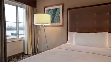 A hotel room with a large bed, a floor lamp, a framed artwork on the wall, and a view of a body of water through the window.