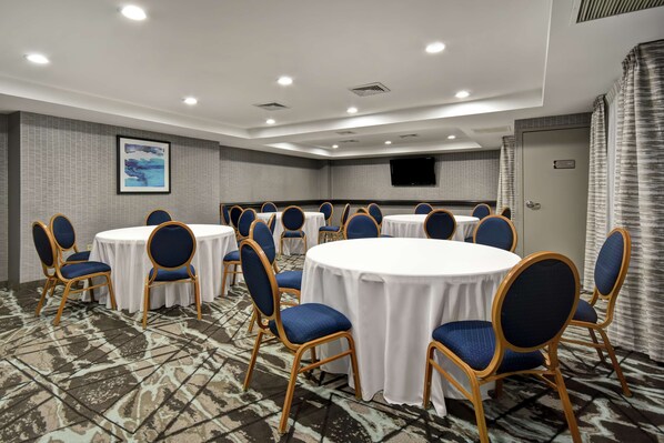 Un salón de conferencias con mesas redondas y sillas azules, una alfombra estampada y un televisor montado en la pared.