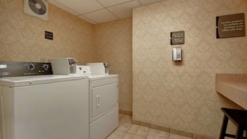 A laundry room with a washing machine, dryer, and a wall-mounted phone. There are signs indicating emergency contact numbers and a note about laundry products available for purchase in the lobby suite shop.