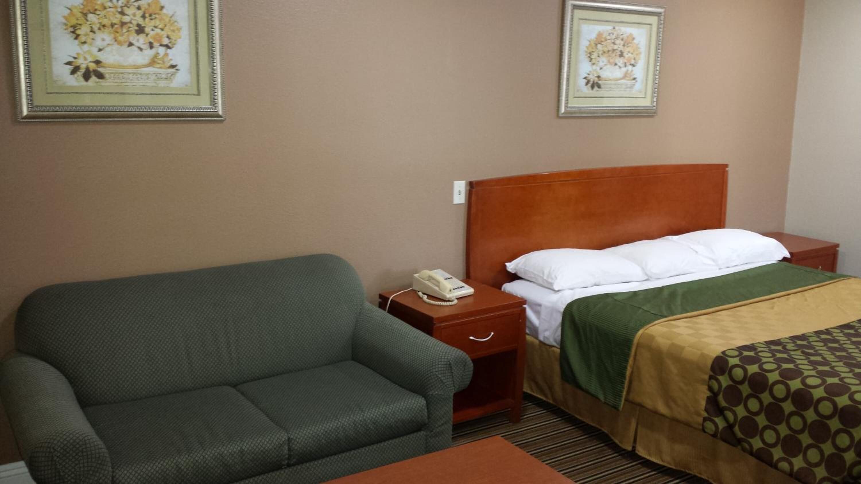 Ein Hotelzimmer mit einem grünen Sofa, einem Bett mit Holz-Kopfteil, einem Telefon auf dem Nachttisch und zwei gerahmten Blumenbildern an der Wand.