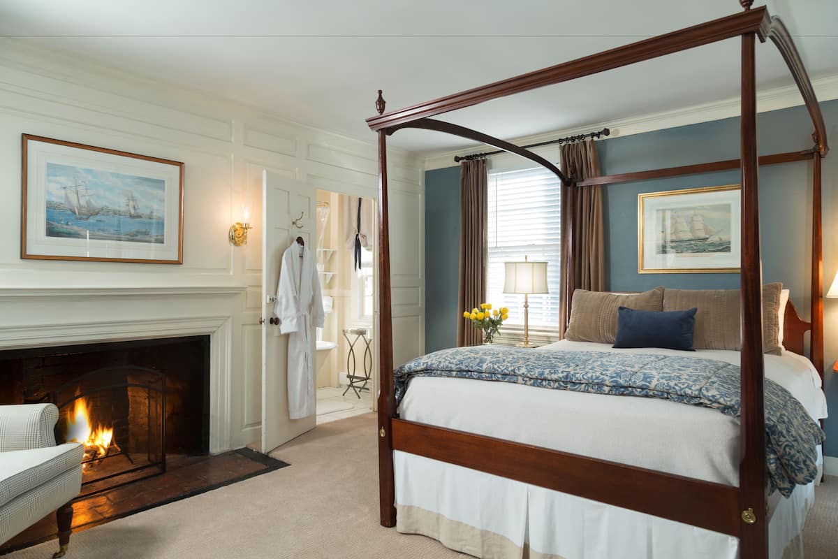 A four-poster bed with a floral bedspread, a fireplace, and a framed seascape painting on the wall.
