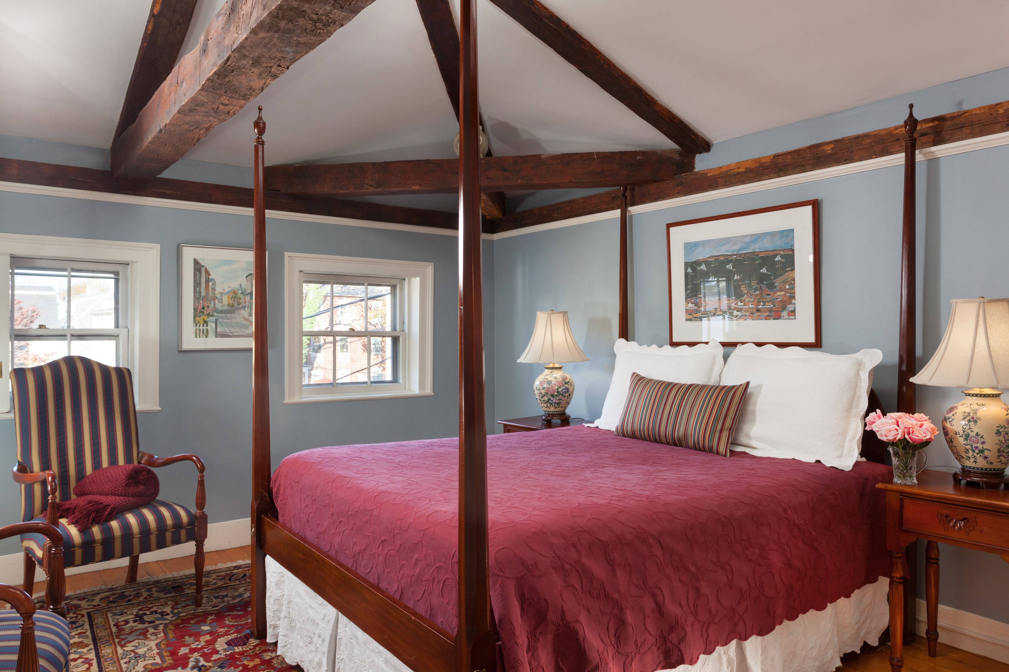 A four-poster bed with a red bedspread, a striped armchair, and a wooden nightstand with a lamp.