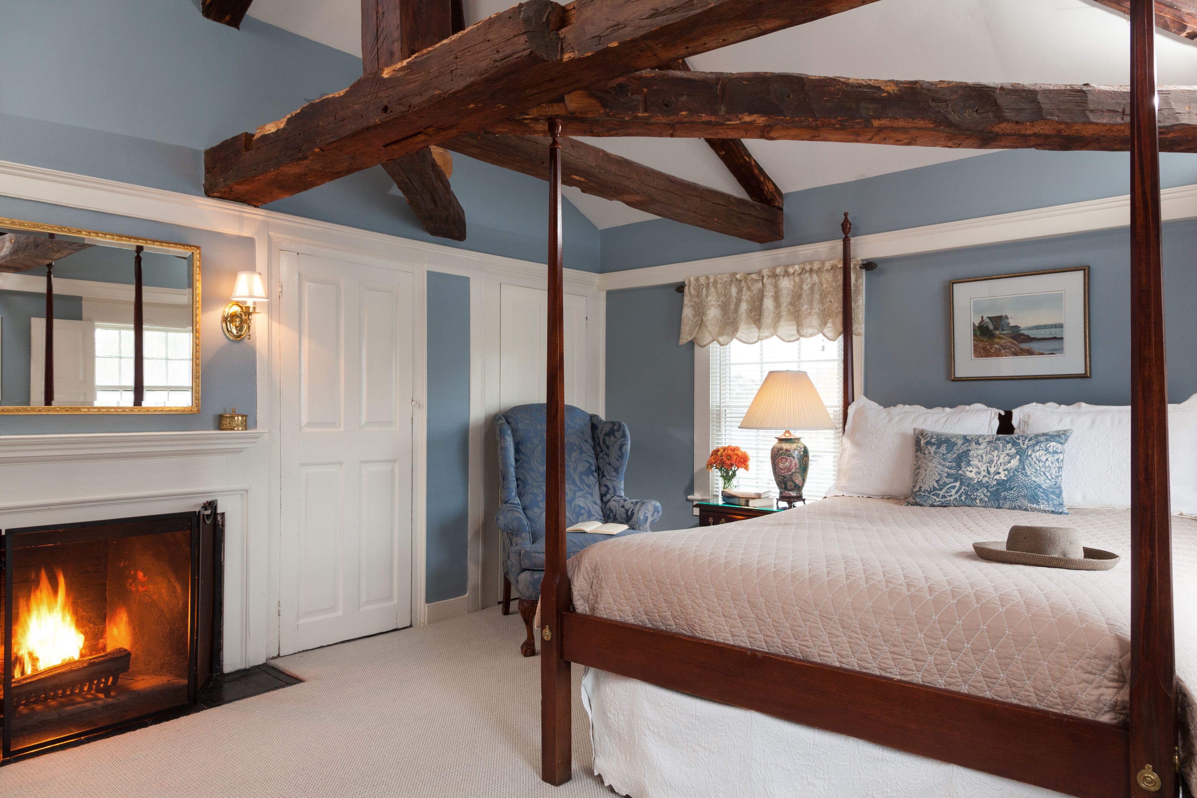A bedroom with a four-poster bed, a fireplace, a blue chair, and a nightstand with a lamp.