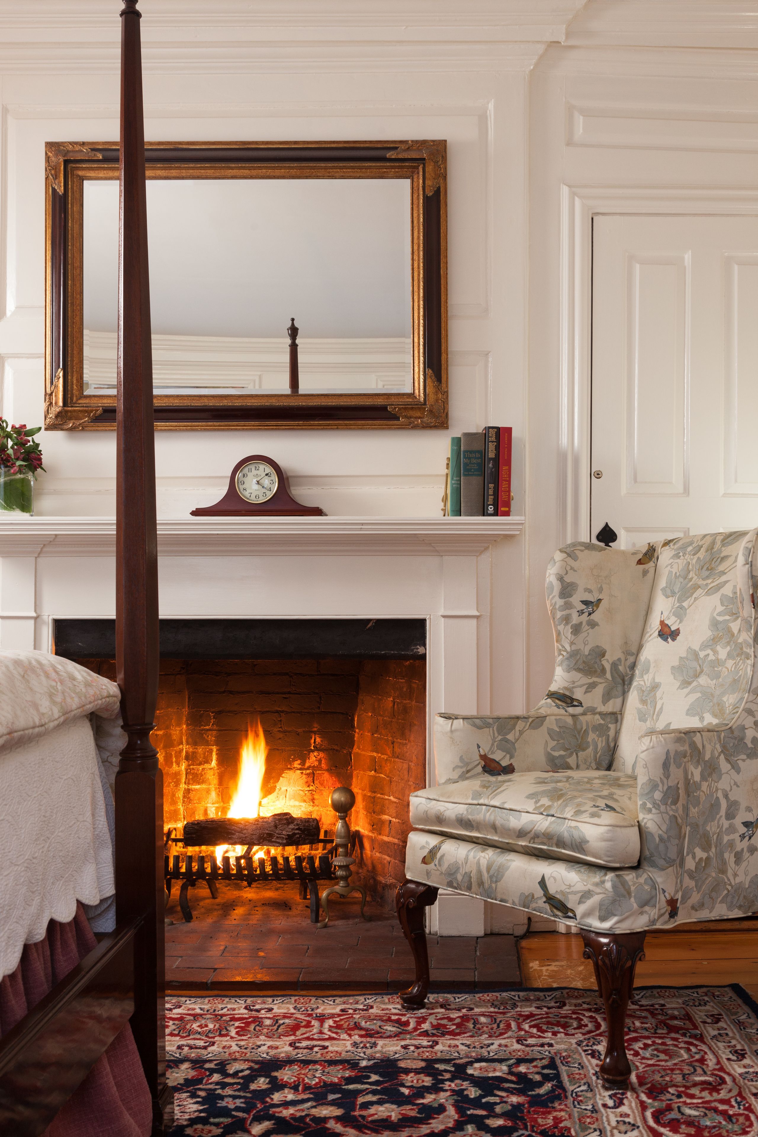 Una habitación tradicional con una cama con dosel, una chimenea encendida, un sillón floral y un espejo grande sobre la chimenea.