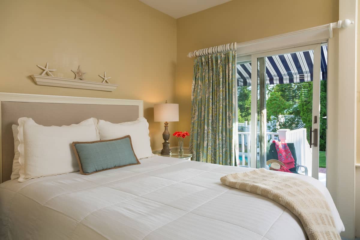A bedroom with a large bed, a nightstand with a lamp, and a view of the outdoors through a sliding glass door.